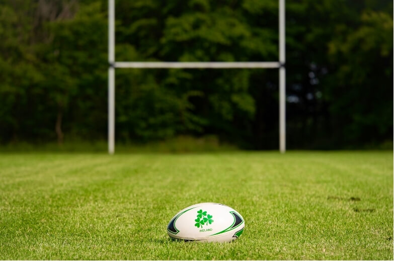 A rugby ball placed on a grassy field with goalposts in the background, symbolising preparation or gameplay in touch football.