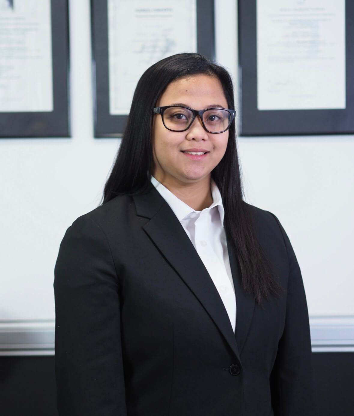 Charisse Castillo, Trainee Practice Manager at M Physio, in a black blazer and white shirt, standing indoors with framed certificates visible behind her.