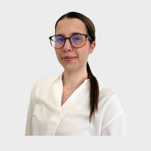 Nicole Bilder, Administrator at M Physio, wearing a white blouse and glasses, smiling softly against a plain light grey background.