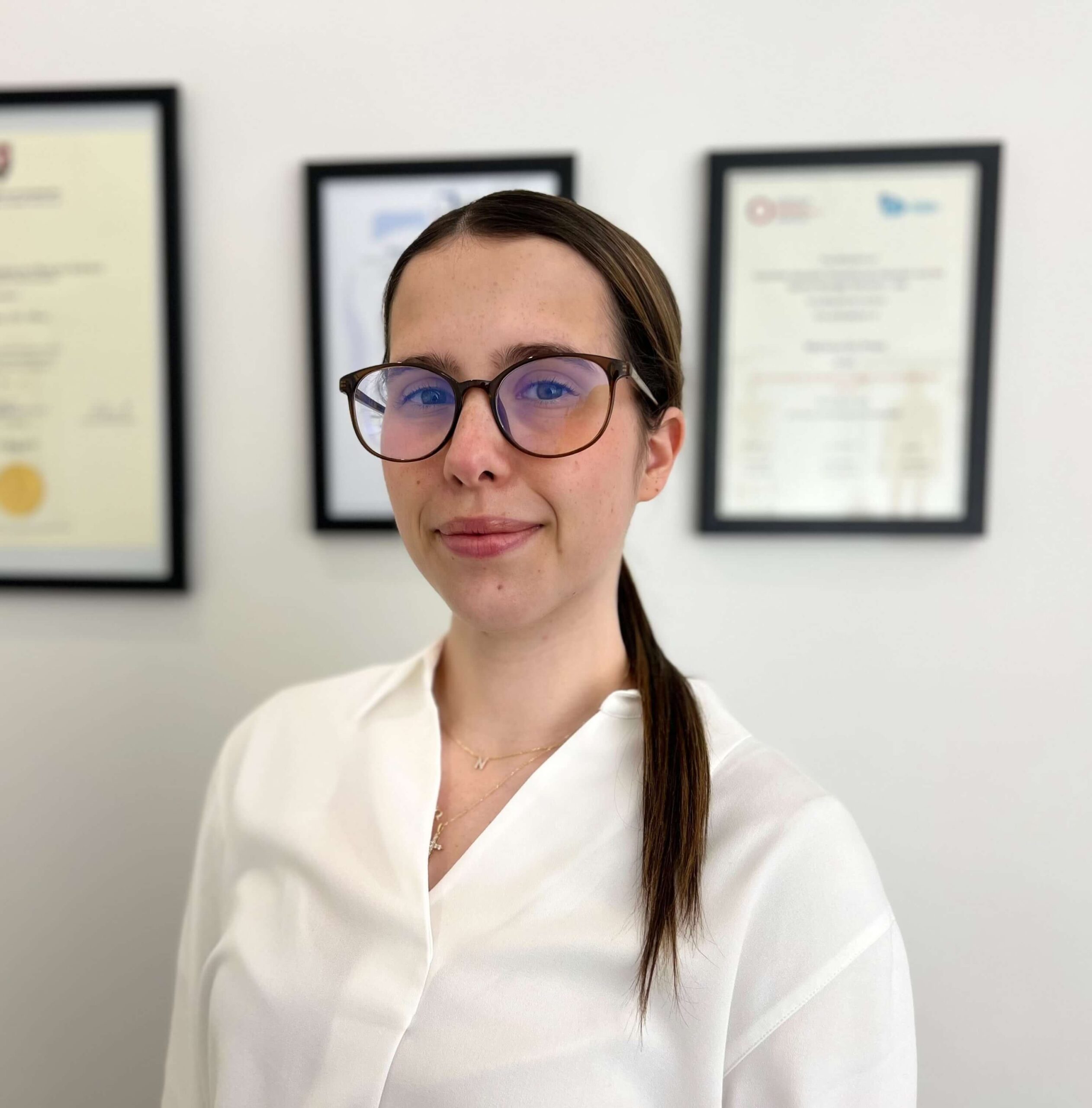 Nicole Bilder, Administrator at M Physio, dressed in a white blouse, standing in front of a wall with framed certificates.