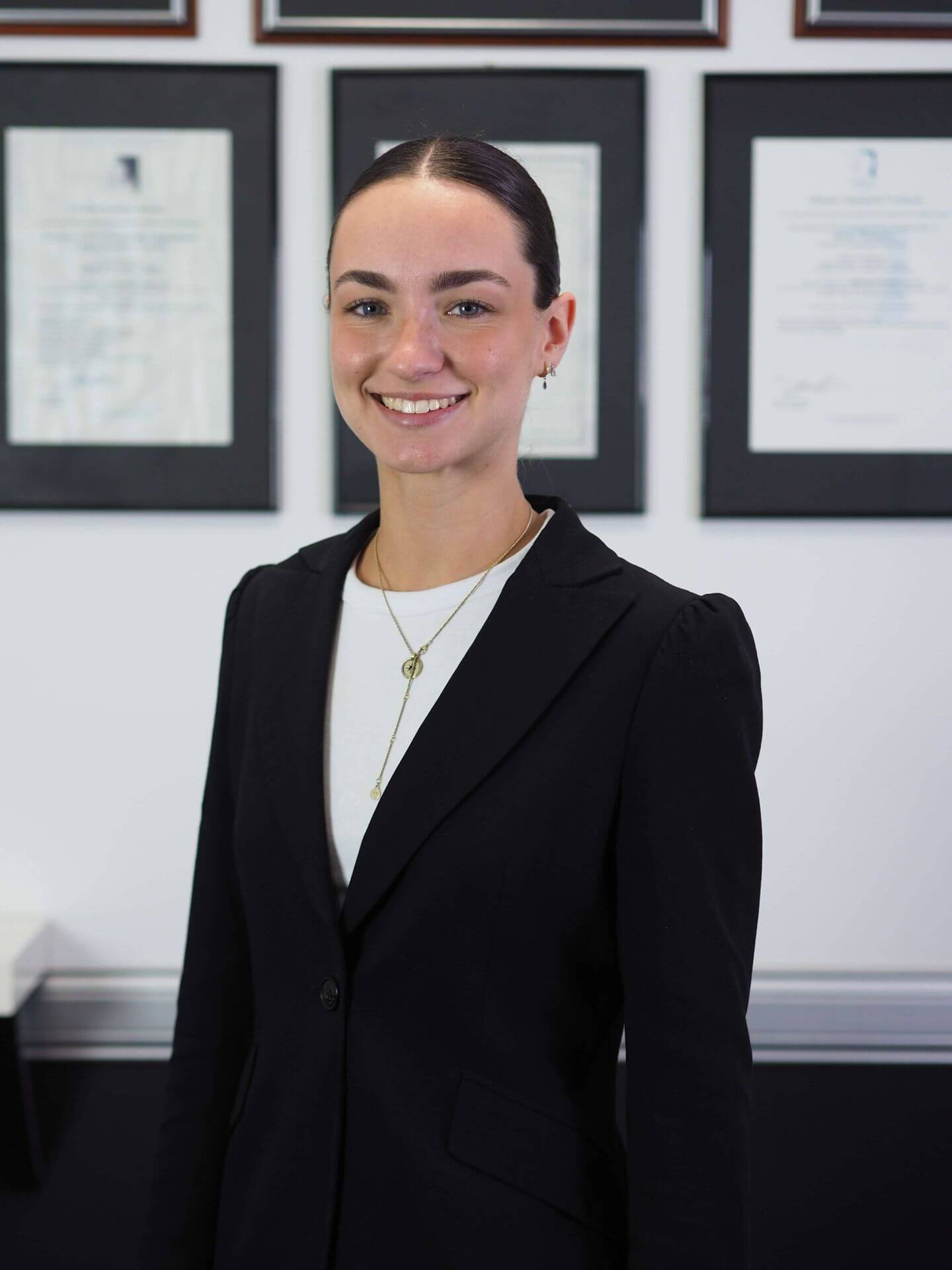 Professional portrait of Rebecca Villegas, Administrator at M Physio, wearing a black blazer and gold necklace, smiling confidently with framed qualifications visible in the background.