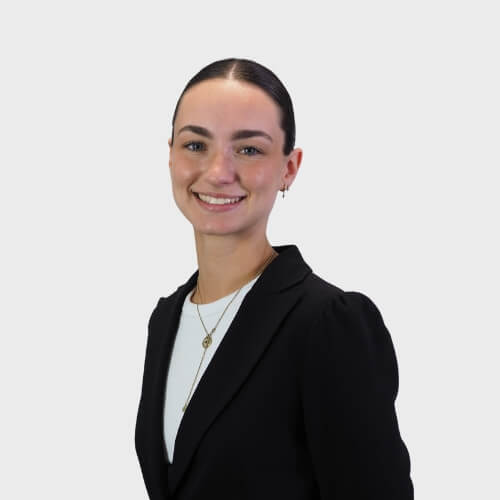 Rebecca Villegas, Administrator at M Physio, smiling in a studio-style headshot against a plain light grey background, dressed in a black blazer and white top with layered gold necklaces.