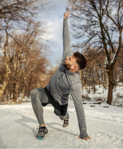 Male athlete performing a dynamic lunge stretch on a snowy trail to prevent cold weather injuries.