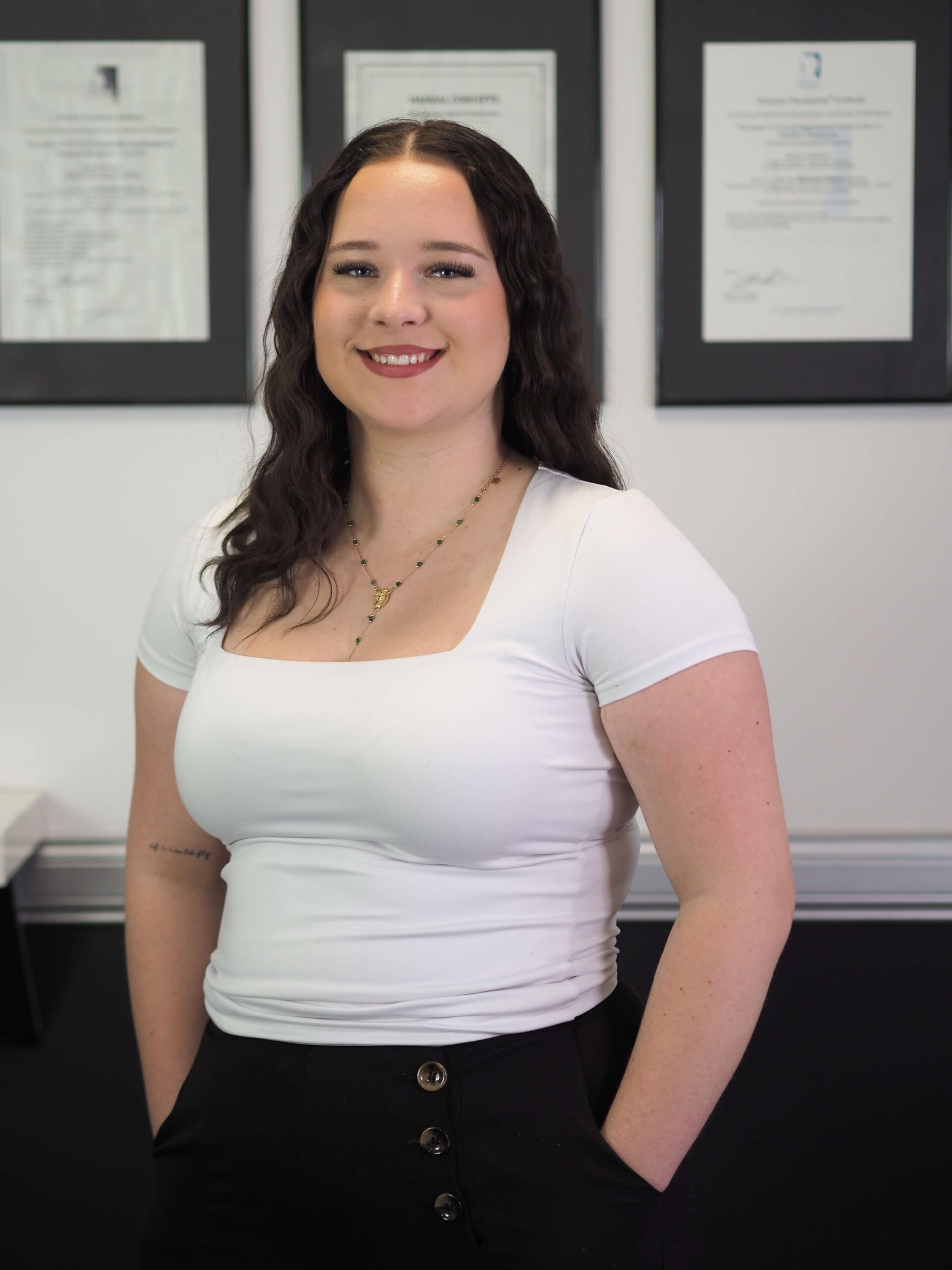 Holly Davies, Administrator M Physio, standing in front of framed certificates, smiling and wearing a white top with black trousers.
