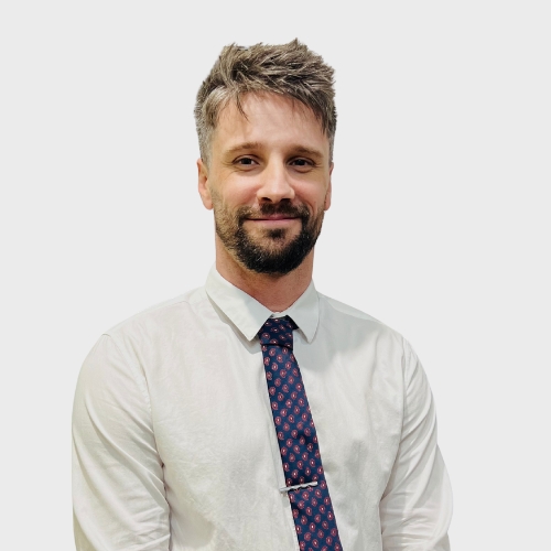 Professional headshot of Joshua Kunde, Administrator, wearing a white shirt and patterned tie, on a plain light grey background.