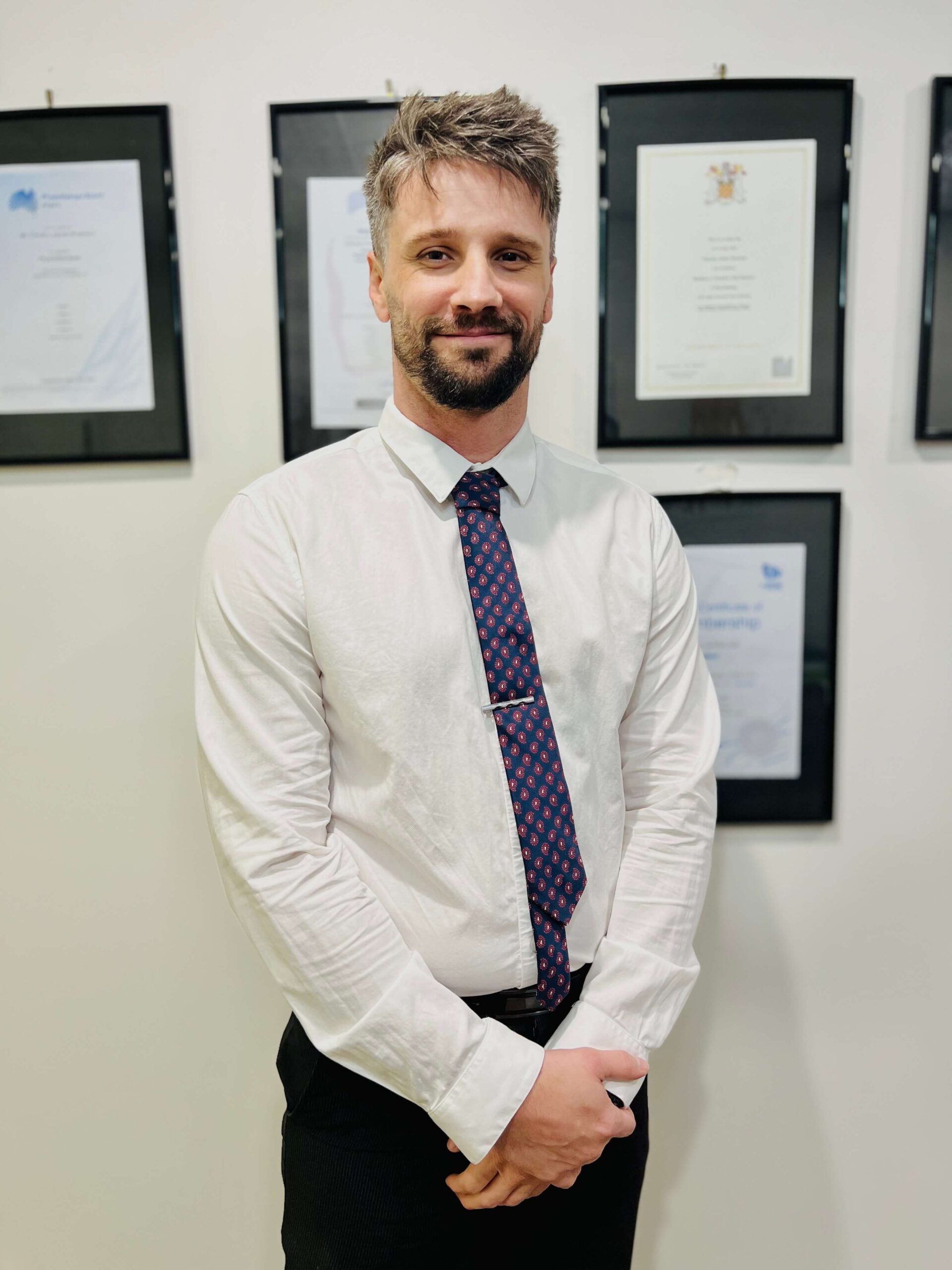 Portrait photo of Joshua Kunde, Administrator, standing in front of framed certificates, wearing a white shirt and patterned tie, smiling at the camera.
