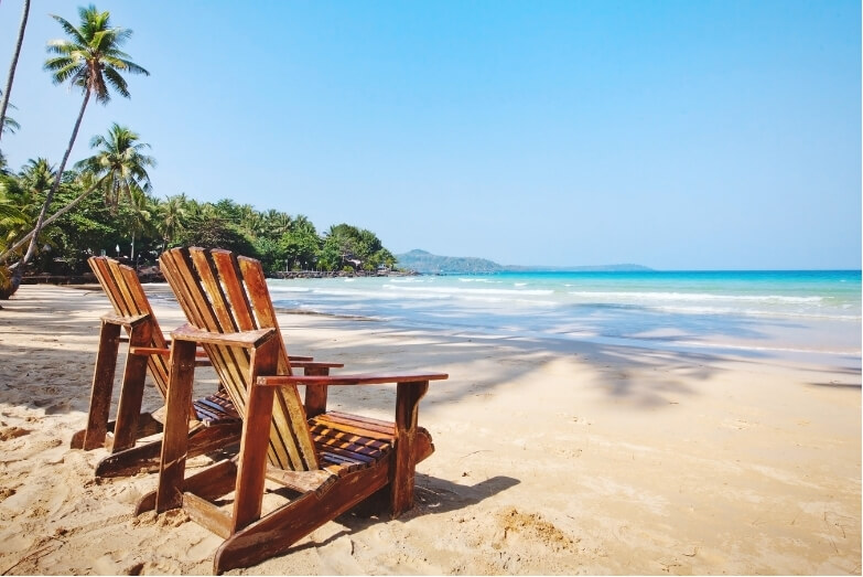 Empty wooden chairs on a sunny tropical beach, symbolising relaxation and the benefits of holiday pain relief.