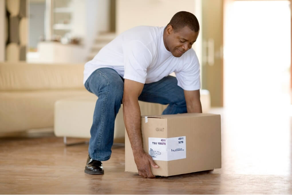 Man using a controlled lifting posture to pick up a cardboard box from the floor.