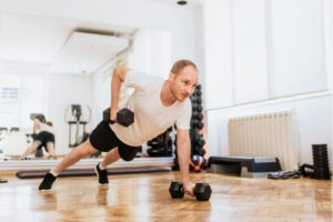 Man performing a single-arm dumbbell row from a plank position while exercising through pain during strength training in a gym.