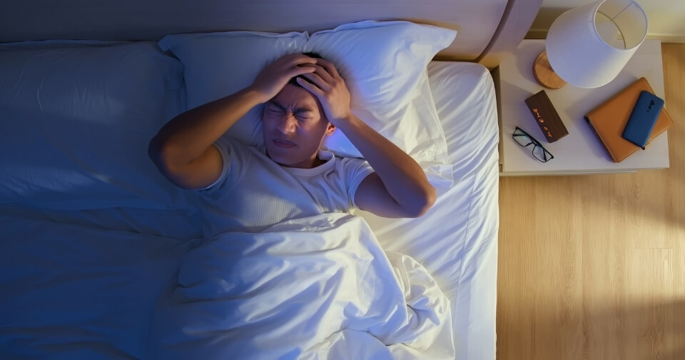 Person lying awake in bed holding their head while experiencing discomfort at night.