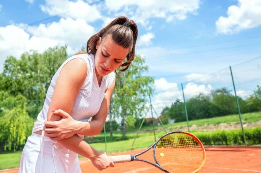 Tennis player holding her painful elbow on an outdoor court during tendon recovery.